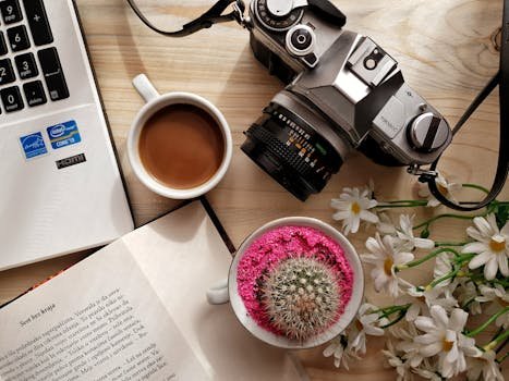 A flat lay of a creative workspace featuring coffee, a vintage camera, book, laptop, and cactus.